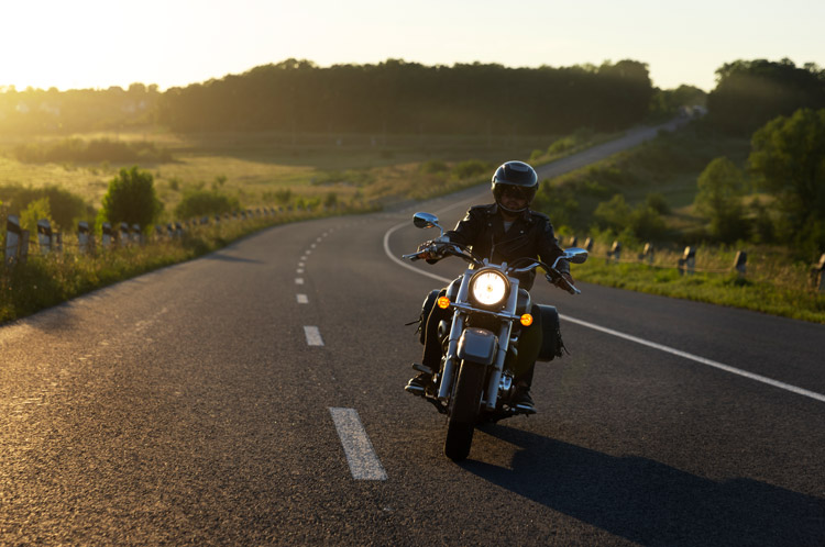 Motard sur une route au coucher du soleil, paysages verdoyants autour.