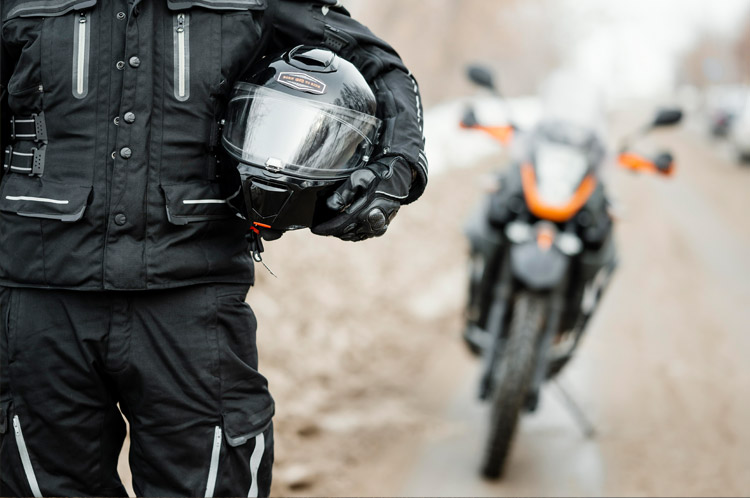 Motard en tenue noire tenant un casque devant une moto floue sur route.